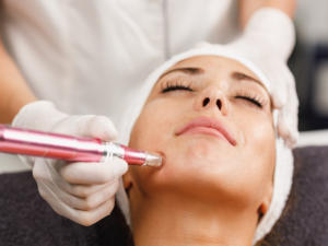 Woman receiving a facial treatment; clinician in gloves uses a pink handheld device on her jawline while she rests with eyes closed.