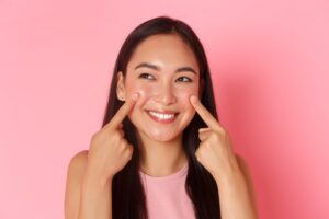Young woman with long dark hair smiling against a pink backdrop, pointing to her cheeks with her index fingers to show skincare glow.