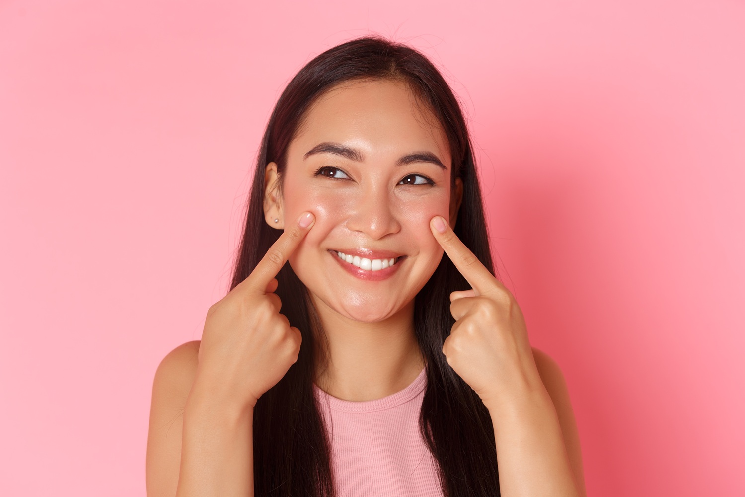 Young woman with long dark hair smiling against a pink backdrop, pointing to her cheeks with her index fingers to show skincare glow.